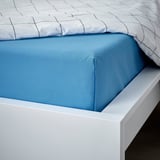 Una cama con una sábana plana DVALA de algodón azul, ligeramente doblada sobre un marco blanco, que promete comodidad y transpirabilidad.