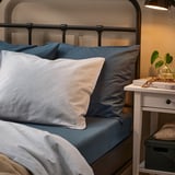 Cama con almohadas a rayas y sólidas de color azul, cabecero de metal negro, mesita de noche blanca con libros y plantas.
