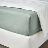 Cama cuidadosamente hecha con sábana de algodón verde grisáceo y almohadas blancas.