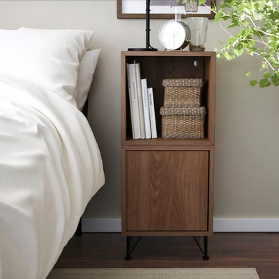 Mesita de noche marrón moderna con almacenamiento abierto y cerrado, al lado de la cama, sostiene libros y cajas.