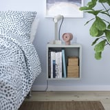 Pequeño estante de cubo blanco EKET, montado al lado de la cama, que contiene libros, reloj rosa, lámpara y vidrio; junto a una gran cama con ropa de cama estampada en azul y blanco.
