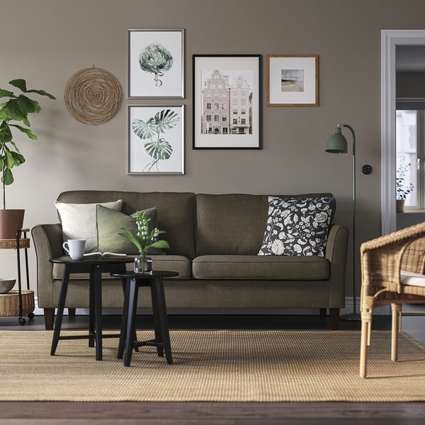 Acogedora sala de estar con sofá EKHOLMA, almohadas verdes, arte de pared de galería, silla de mimbre y mesa auxiliar con taza y planta.