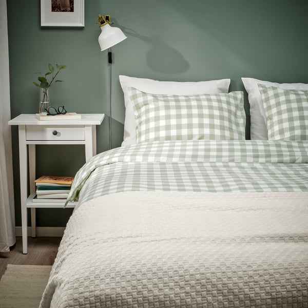 Dormitorio con edredón de cuadros gris-verde y almohadas sobre cama blanca, mesita de noche con lámpara y libros, pared verde.