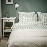 Dormitorio con edredón de cuadros gris-verde y almohadas sobre cama blanca, mesita de noche con lámpara y libros, pared verde.