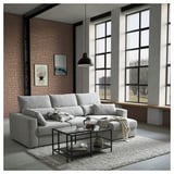 Moderno salón comedor con sofá ESKILSTUNA gris claro, pared de ladrillo, grandes ventanales.