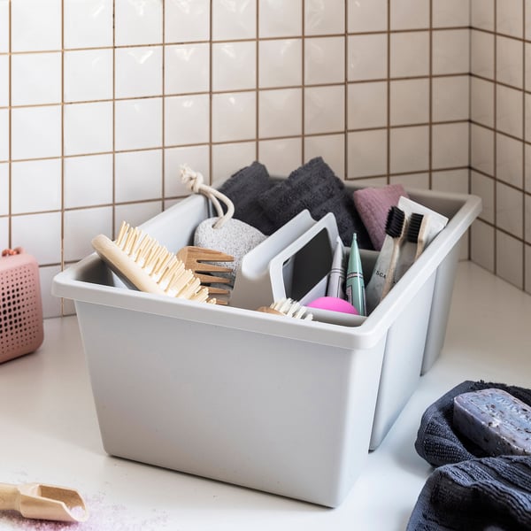 El contenedor de almacenamiento de plástico blanco con múltiples compartimentos contiene artículos de baño, incluidos cepillos y artículos de tocador, organizados en una encimera de azulejos.
