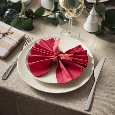 Un juego de mesa con un plato blanco, una servilleta roja doblada, un tenedor, un cuchillo, una copa de vino, hojas verdes y un pequeño regalo.