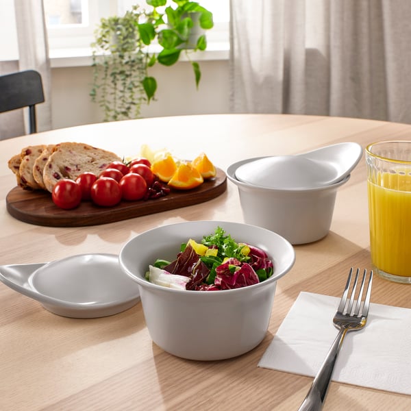 Mesa de comedor de madera con cuencos, pan, frutas y zumo. El tazón central tiene ensalada colorida, rodeada de tenedores, servilletas y tazones blancos cubiertos.