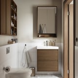 Baño moderno con mueble de madera, espejos de luces, lavabo blanco, toalla para colgar e inodoro.