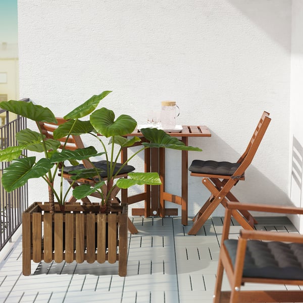 Escena de balcón con mesa de madera, silla y gran planta verde FEJKA en jardinera de madera.