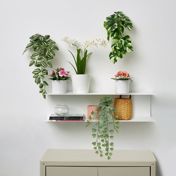 Estanterías blancas con diversas plantas artificiales en macetas de colores, algunas en cascada, contra una pared de luz.