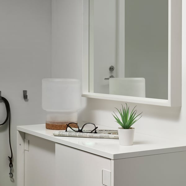 Baño moderno con encimera blanca, pequeño lavabo de cerámica blanca, espejo de seguridad y diminuta planta verde artificial FEJKA.