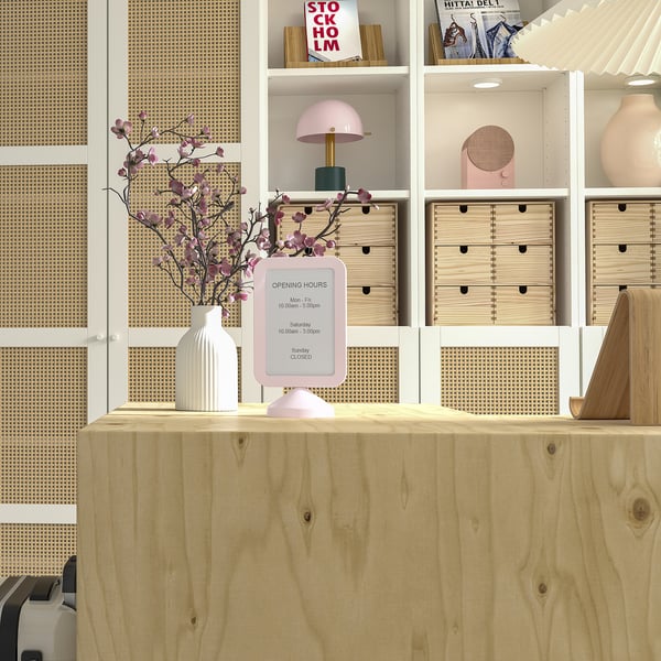 Marco de fotos rosa de doble cara, FIKONTRÄD, sobre escritorio de madera, rodeado de estanterías con libros y decoración, destacando su pequeño tamaño.