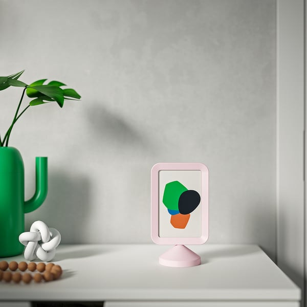 Un marco de fotos rosa en un estante blanco que muestra arte colorido. A su lado, un jarrón de cactus verde con una planta y una pequeña escultura blanca.