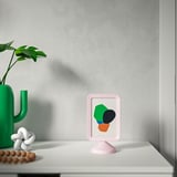 Un marco de fotos rosa en un estante blanco que muestra arte colorido. A su lado, un jarrón de cactus verde con una planta y una pequeña escultura blanca.