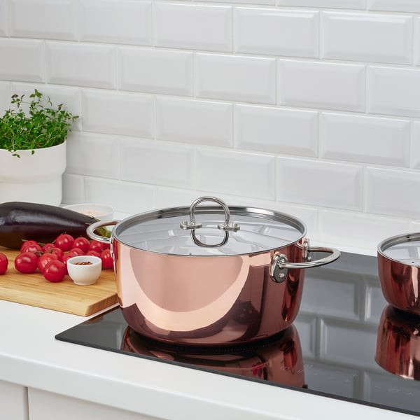 Una cocina moderna con ollas de acero inoxidable y cobre, tomates y una berenjena en una tabla de cortar de madera junto a una hierba blanca en maceta.