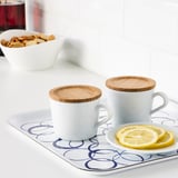 Dos tazas blancas con tapas de bambú en una bandeja con rodajas de limón, demostrando su uso como fundas para tazas.