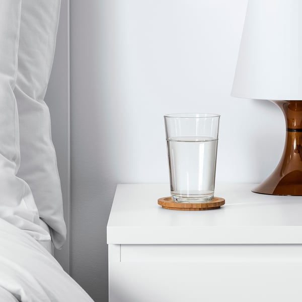 Vaso transparente con agua en mesita de noche blanca al lado de la cama, cubierta por una tapa redonda de bambú.