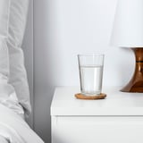 Vaso transparente con agua en mesita de noche blanca al lado de la cama, cubierta por una tapa redonda de bambú.