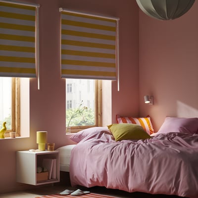 Acogedor dormitorio con paredes rosas, una cama grande cubierta con ropa de cama de color púrpura claro y cortinas a rayas sobre dos ventanas, con una decoración moderna.