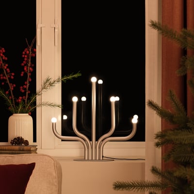 Candelabro led metálico encendido en el alféizar de la ventana, emitiendo una luz cálida, rodeado de ramas de pino y bayas.