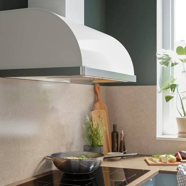 Cocina moderna con campana extractora blanca, estufa y encimera de madera; vegetación, sartenes y manzanas cerca.