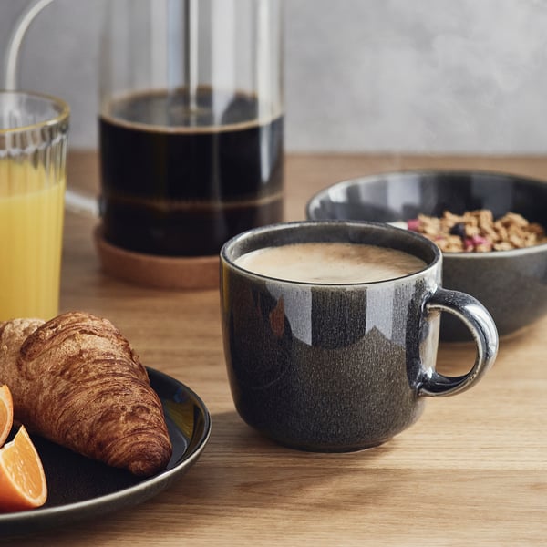 Taza gris oscuro, jugo de naranja, croissant y granola en una mesa de madera, mostrando una elegante configuración de desayuno.