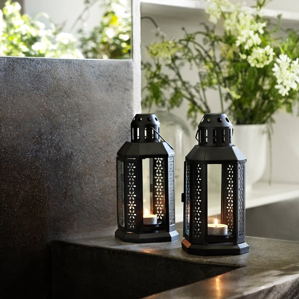 Dos faroles GLIMMA negros con velas encendidas sobre una superficie de piedra. Una linterna tiene una vela candelita. Diseño decorativo y metálico con aberturas.
