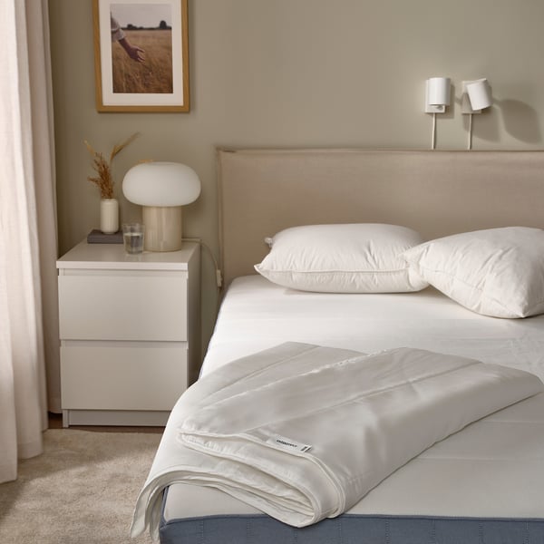 Cama pulcra con edredón GRÄSROTSFLY, lyocell sedoso, cabecero beige. Al lado del cajón hay una lámpara, un jarrón con plantas secas. Decoración minimalista.