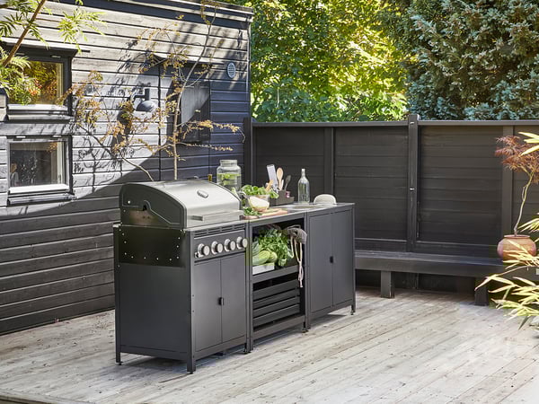 Configuración de cocina al aire libre con parrilla GRILLSKÄR negra, fregadero y unidades de almacenamiento en cubierta de madera, rodeada por una valla de madera oscura.
