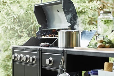 Cocina al aire libre con parrilla a gas y quemador lateral; humo que se eleva, ollas de acero inoxidable, fondo verde.