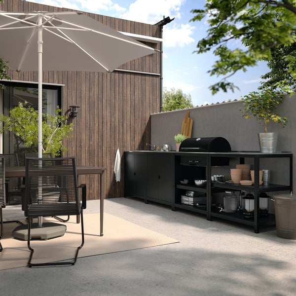 Moderna cocina al aire libre. Mueble GRILLSKÄR negro con parrilla, fregadero y estantes. Silla de madera, sombrilla grande y plantas. Casa al fondo.