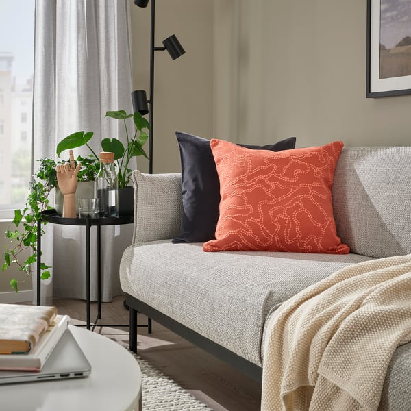Salón moderno con sofá gris, cojín GULDFLY naranja, suelo de madera y planta interior. El cojín es cuadrado y estampado.
