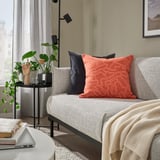 Salón moderno con sofá gris, cojín GULDFLY naranja, suelo de madera y planta interior. El cojín es cuadrado y estampado.