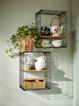 El estante de pared de alambre negro y madera sostiene platos, tazones y plantas cerca de la ventana.