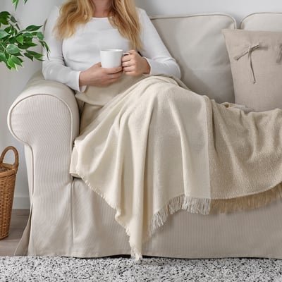 Persona vestida de blanco en el sofá, sosteniendo una taza, debajo de la manta de flecos, cerca de la planta.