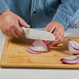 Una persona corta cebolla morada en rodajas en una tabla de cortar de madera con un cuchillo de cerámica. Las cebollas están parcialmente picadas, mostrando el filo de los cuchillos.