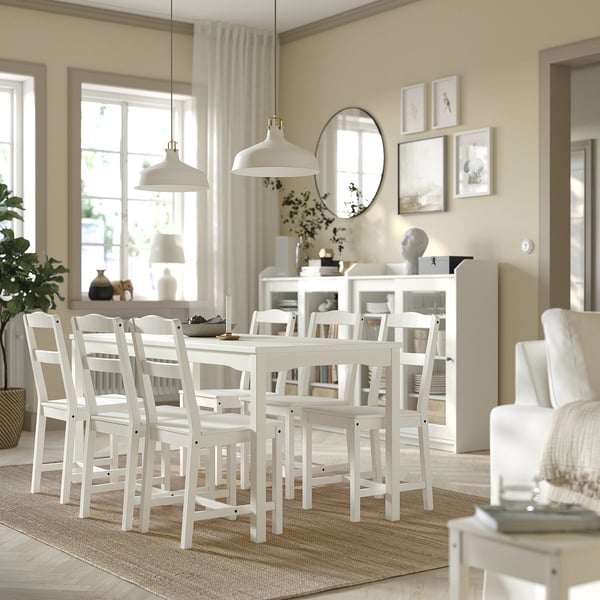 Comedor luminoso con mesa y sillas de madera blanca, luces colgantes, gran ventanal y alfombra estampada.