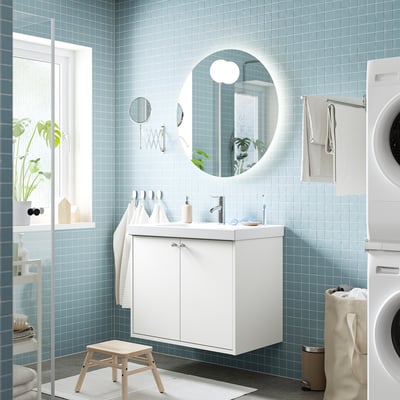 Baño moderno con tocador HAVBÄCK blanco flotante, espejo redondo, azulejos azules, plantas, toallas, lavadora, taburete de madera.