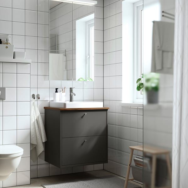 Baño moderno con tocador HAVBÄCK gris oscuro. Encimera de madera, lavabo blanco, espejo grande. La toalla cuelga a la izquierda, el taburete a la derecha.