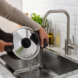 Una persona vierte agua de un colador sobre un fregadero en una cocina, usando la tapa HEMLAGAD.