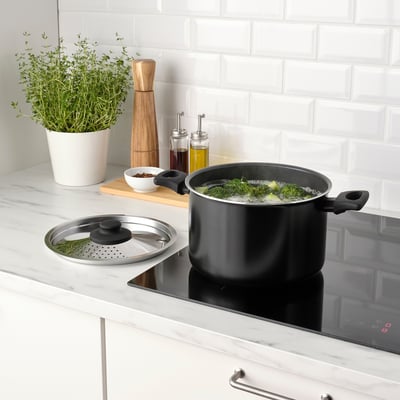 Utensilios de cocina negros con tapa, brócoli hirviendo. Encimera de cocina con hierbas, condimentos y estufa digital.
