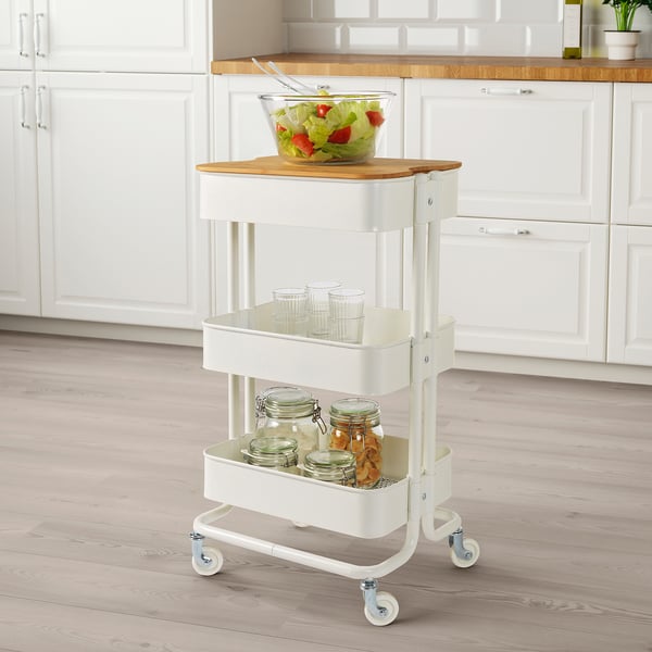 Carro de cocina blanco con ruedas, sosteniendo un tazón de vidrio con ensalada en una tabla de cortar de bambú, pilas de tazas y frascos con bocadillos.