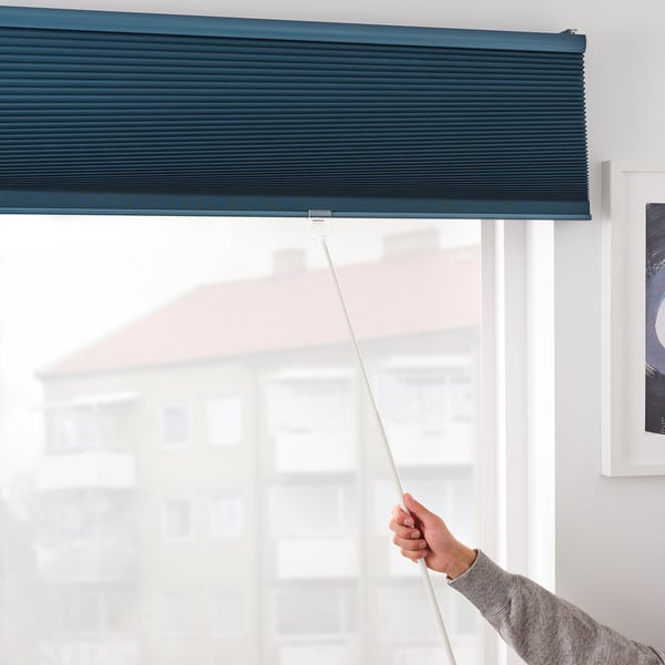 Una mano ajusta una persiana azul oscuro que oscurece la habitación, controlando la luz y proporcionando privacidad. La persiana tiene un cordón oculto y seguro para la protección de los niños.