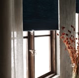 Una ventana con persianas grises que oscurecen la habitación HOPPVALS, marco de madera y flores secas en el costado.