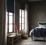 Acogedor dormitorio con persianas azul oscuro, cortinas claras, banco con libros y flores secas, cama con manta azul oscuro, iluminación tenue.