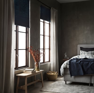 Dormitorio luminoso con grandes ventanales, madera clara, cortinas oscuras. Jarrón con flores secas en banco, cama gris y azul.