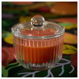 El frasco de vidrio transparente con lados estriados sostiene una vela naranja, cubierta con una tapa plateada. Se coloca sobre una tela colorida y estampada.
