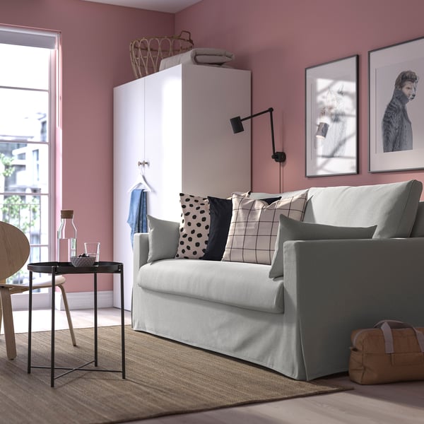 Acogedora sala de estar con sofá cama gris, paredes rosas, almohadas estampadas, mesa auxiliar con jarrones y armario blanco.