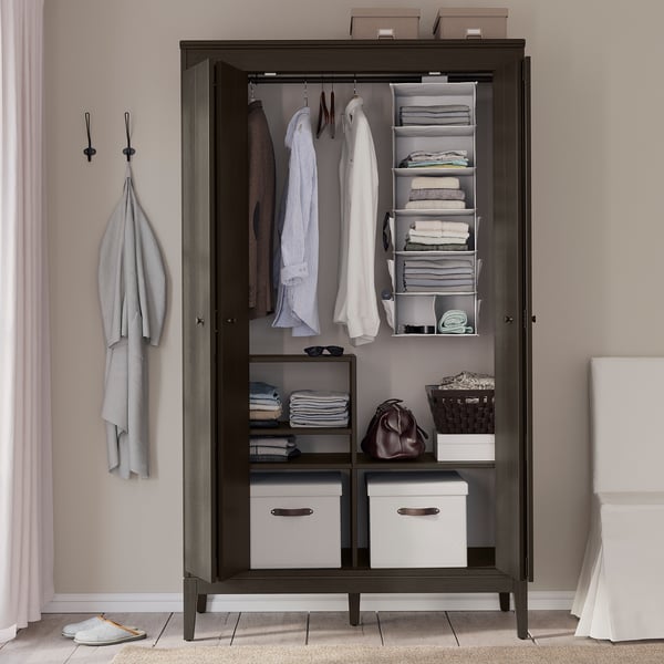 Armario de madera oscura de IDANÄS con puertas transparentes, ropa organizada, perchas y cajas de almacenamiento.